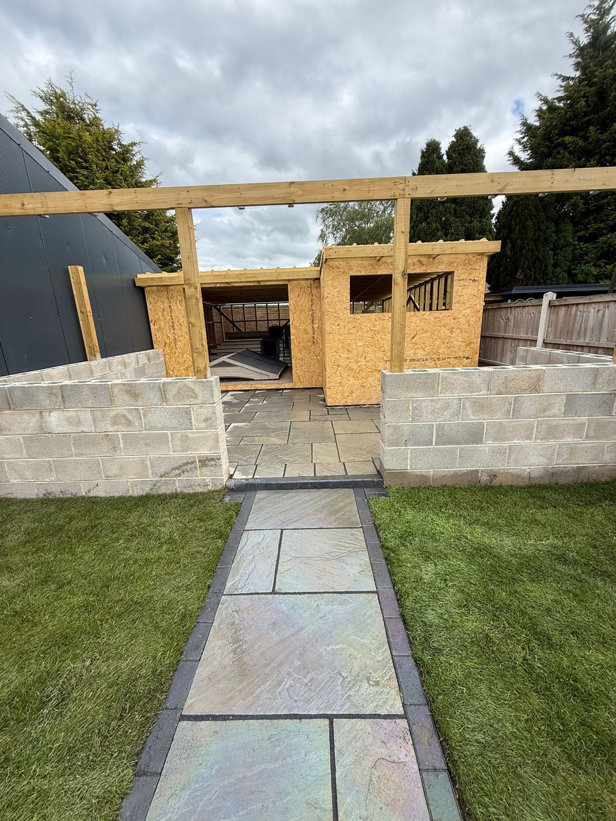 Stone path leading to a small unfinished wooden shed behind a low cinderblock wall and lawn.