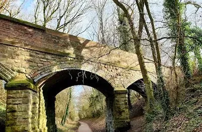 Stone arch bridge over a path lined with trees; overcast sky.