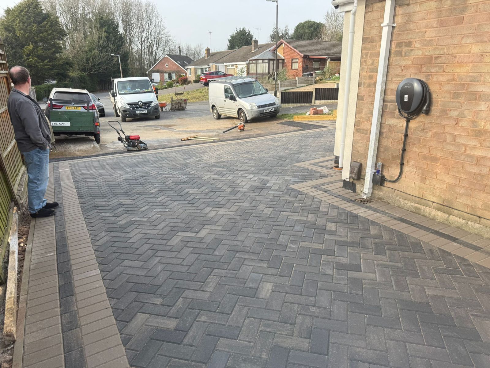 A person stands on a dark gray herringbone-patterned paved driveway next to a brick house with an EV charger on the wall.