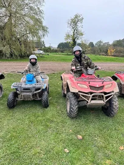 Two people on ATVs in a grassy field, wearing helmets. One blue ATV, one red ATV. Cloudy day.
