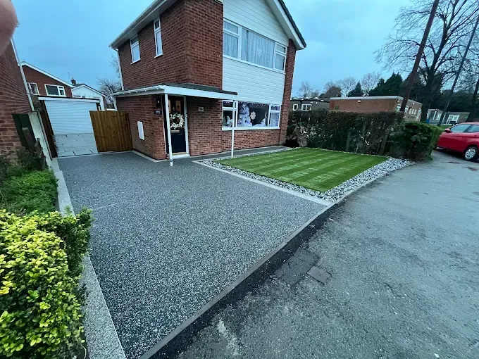 Grey resin driveway with light-colored buildings, dark car, and a wall in the background.