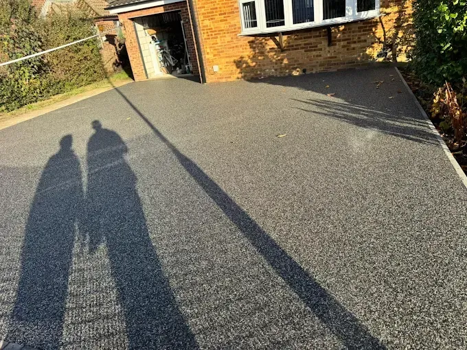 A two-story beige house with a resin driveway and green lawn, surrounded by trees.