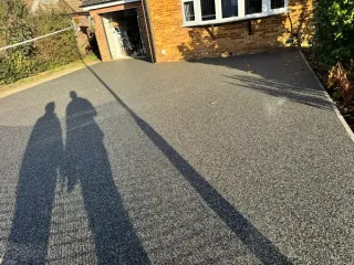 A two-story beige house with a resin driveway and green lawn, surrounded by trees.
