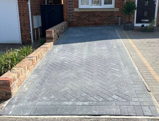 Gravel driveway in front of a brick house with windows. Red fence and a flowerbed on the sides.