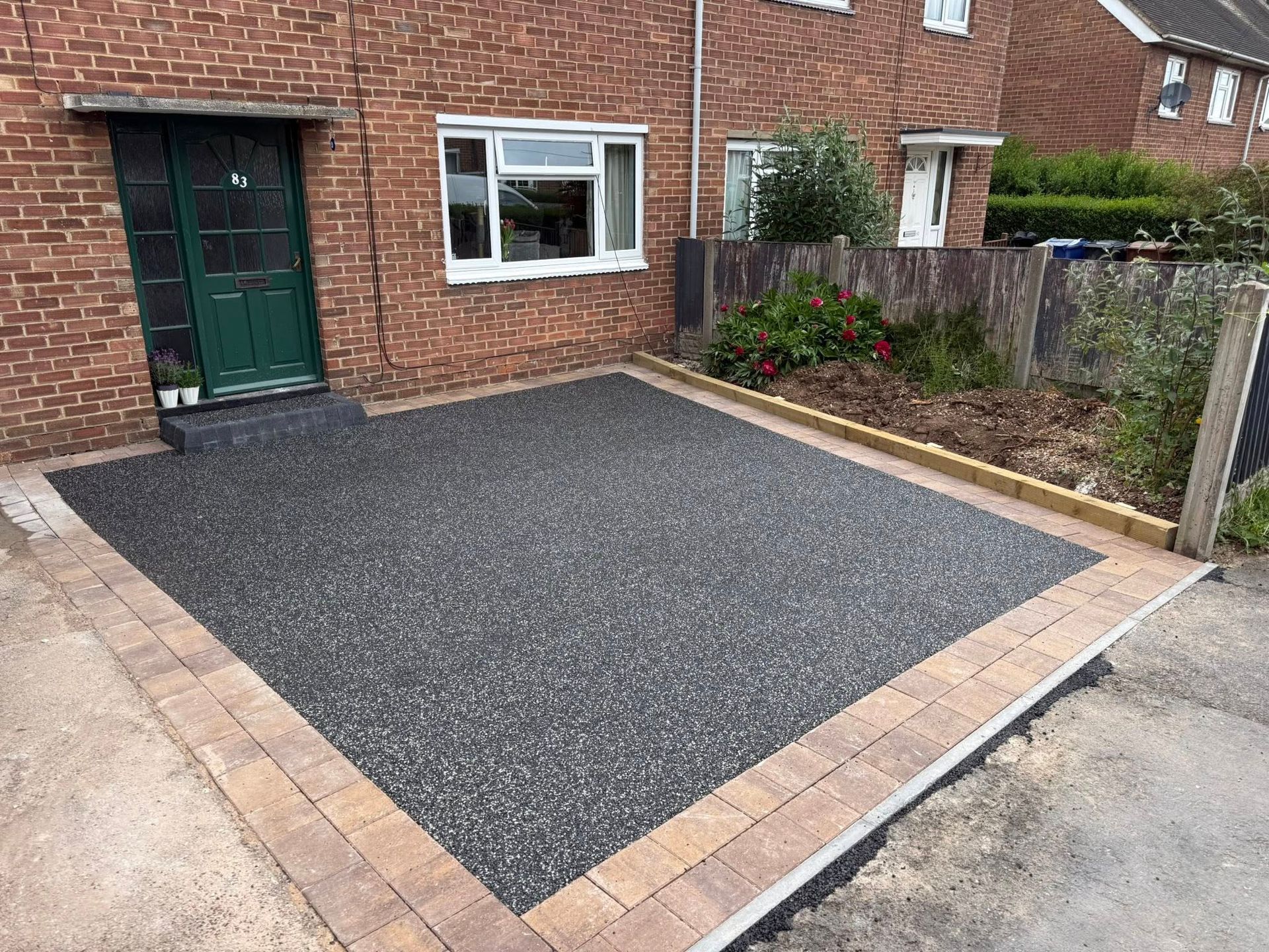 Gravel front driveway bordered by brick pavers beside a brick house and garden fence.
