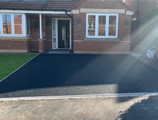 Gravel driveway in front of a brick house with windows. Red fence and a flowerbed on the sides.