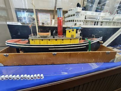 Model tugboat, yellow and black, in a wooden box, with a large white ship in the background.
