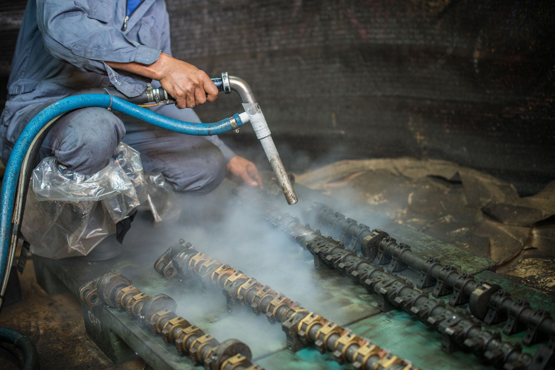 Person in blue coveralls using a pressure washer to clean metal machine parts in an industrial setting.
