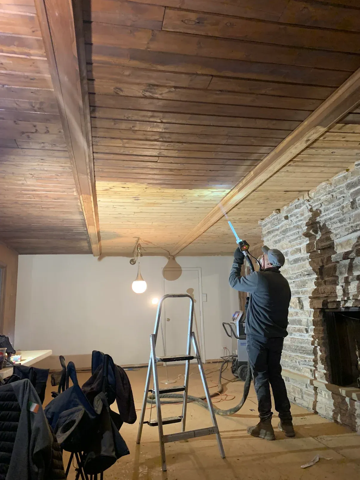 Man spraying ceiling beams, standing on a ladder in a room with a stone fireplace.