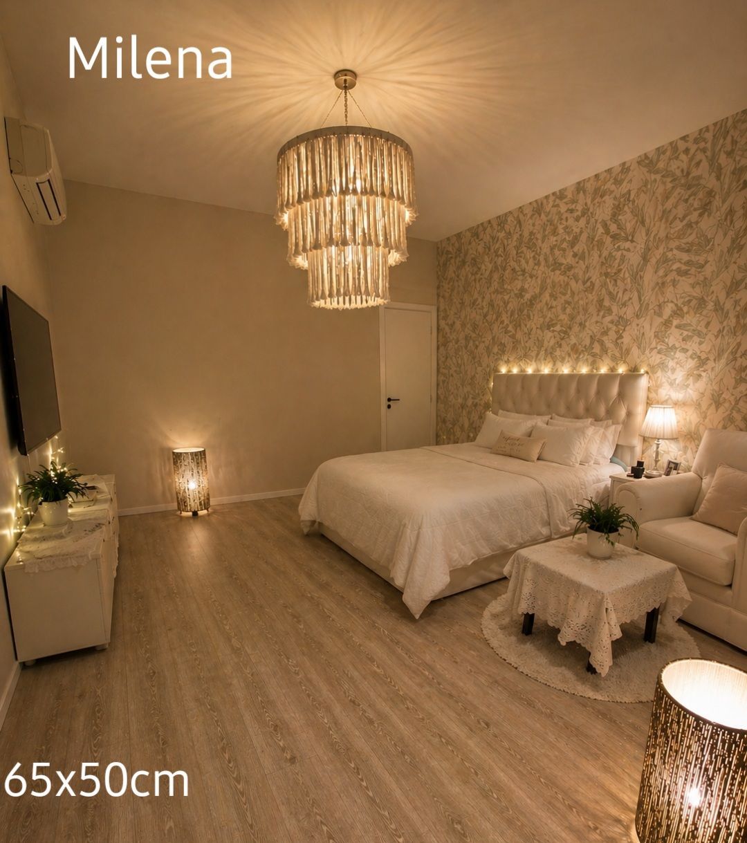 Acogedor dormitorio beige con lámpara de araña, pared decorativa estampada, cama blanca y luz cálida y suave.