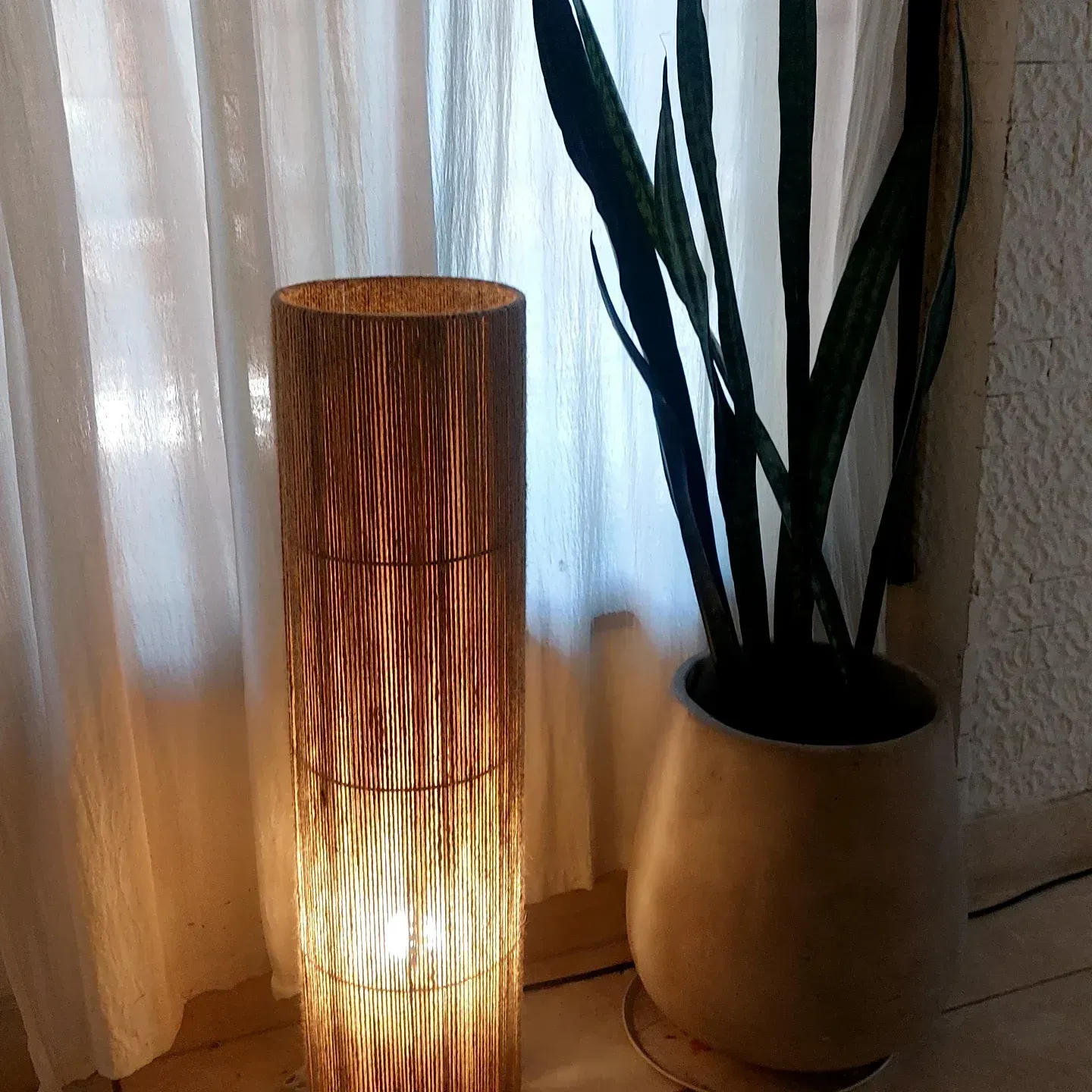 Lámpara de pie de luz cálida junto a una planta en maceta junto a una ventana con cortinas.