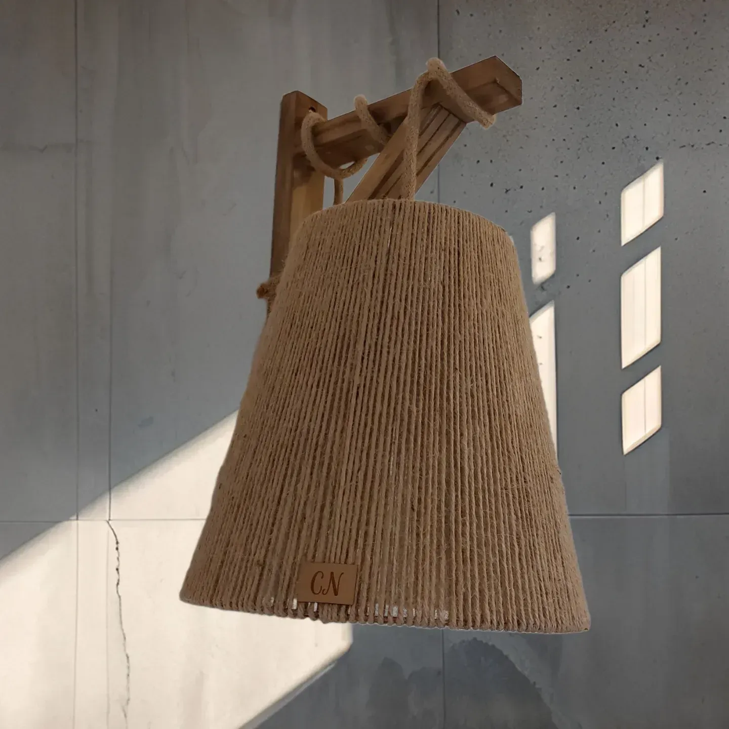 Lámpara de pared de mimbre tejido con soporte de madera que proyecta luz y sombra sobre una pared de hormigón.