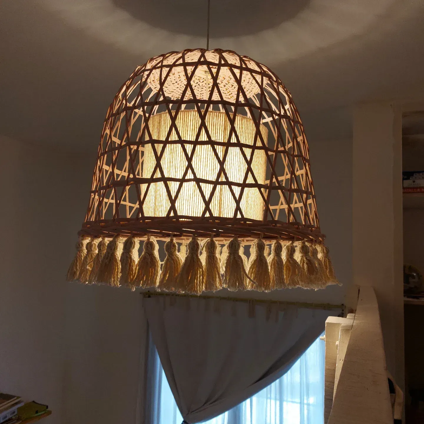 Lámpara colgante de ratán tejido con borlas, que emite una luz cálida sobre una ventana con cortinas en una habitación.