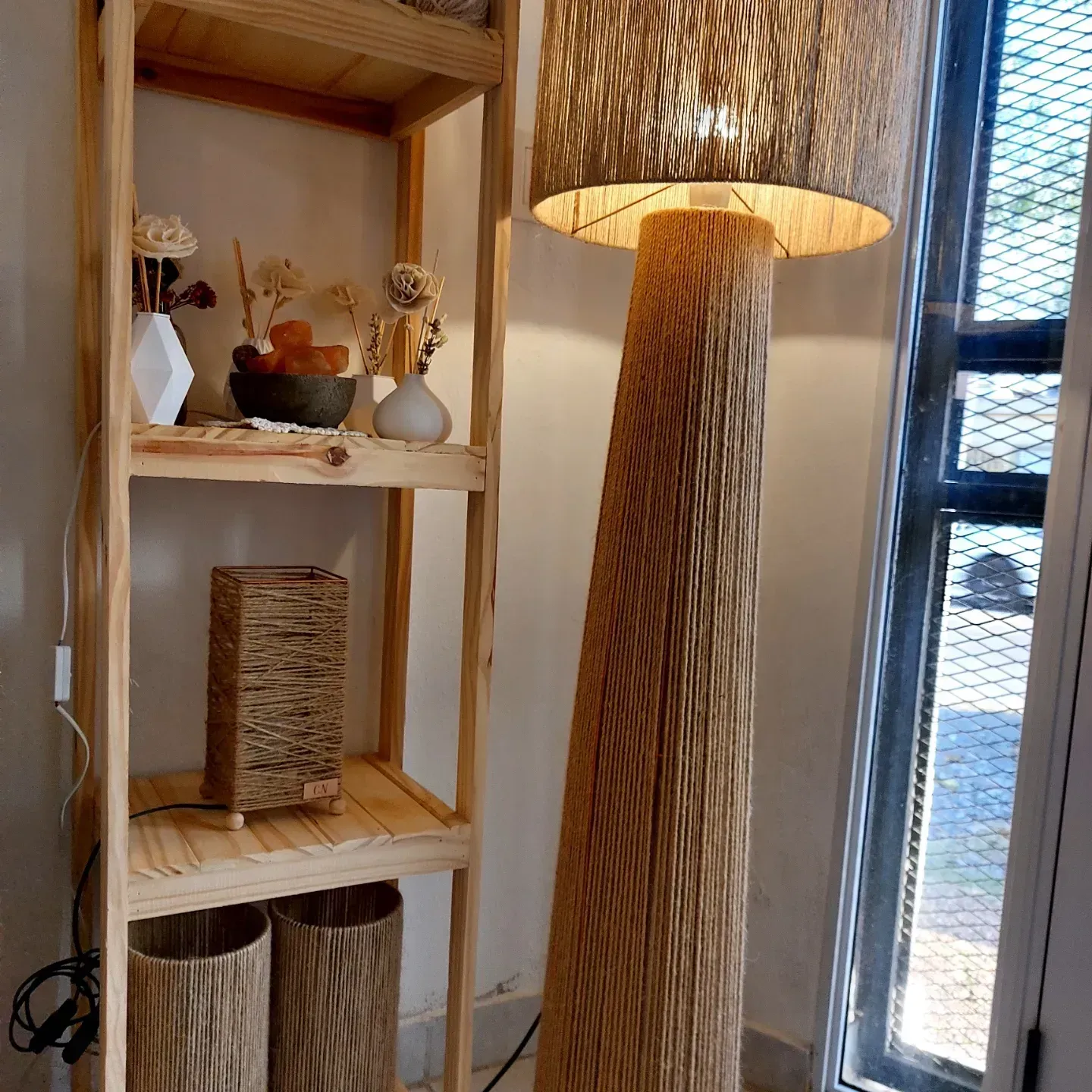 Estantería de madera con cestas tejidas y elementos decorativos junto a una lámpara de pie alta tejida cerca de una ventana.