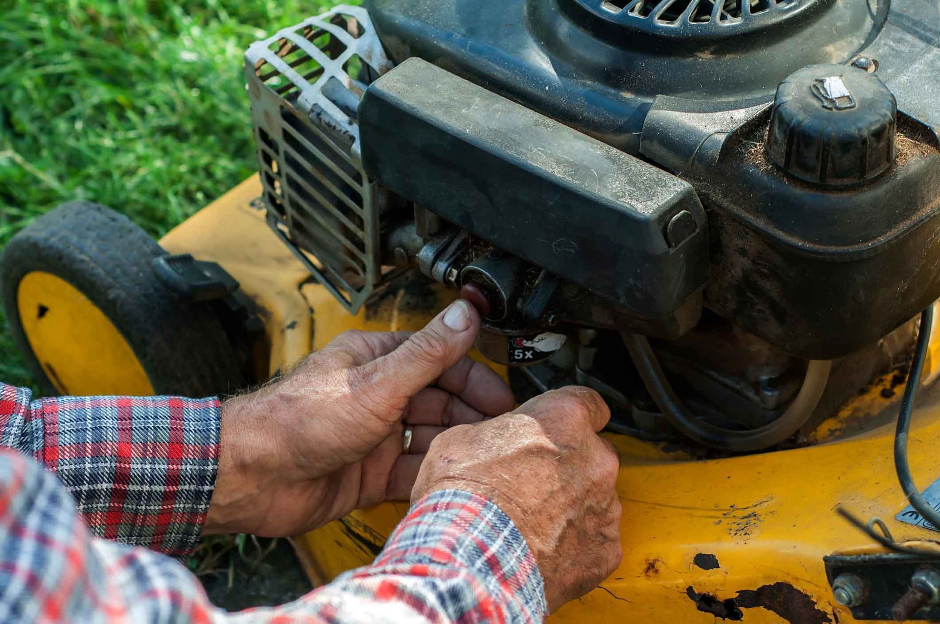 A person pressing the red primer bulb on a yellow lawnmower engine.