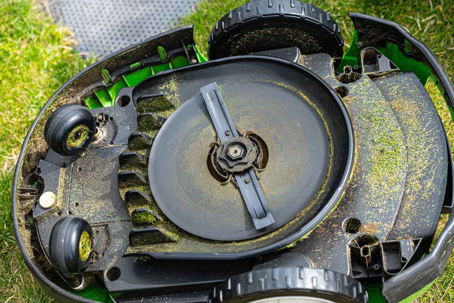 Underside of a robotic lawnmower on grass, showing the central rotating cutting blade and small wheels.