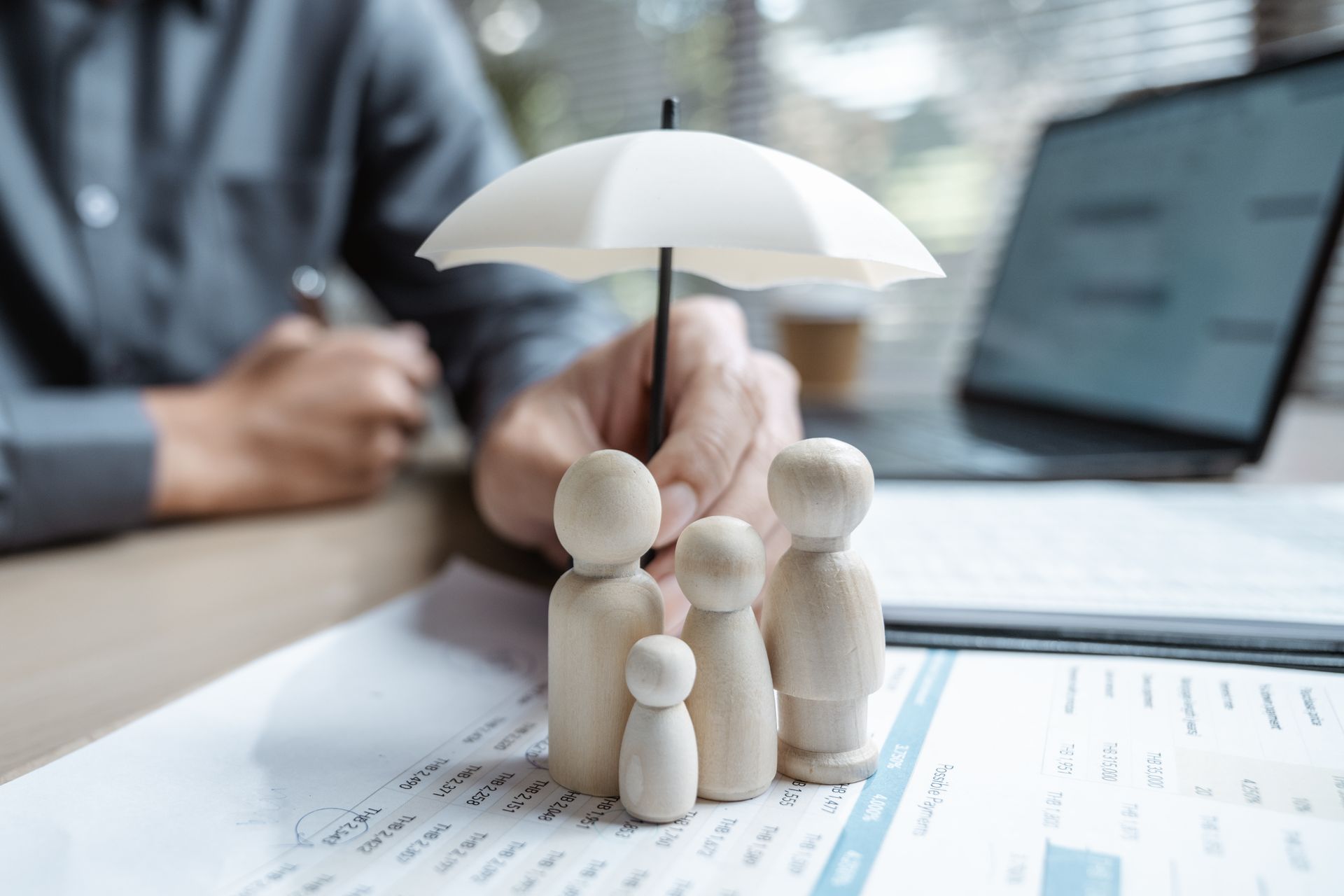 Person holding a miniature umbrella over wooden family figurines, symbolizing insurance coverage.