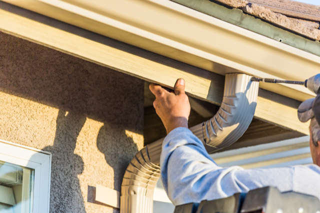 A man is fixing a gutter on a house with a drill.