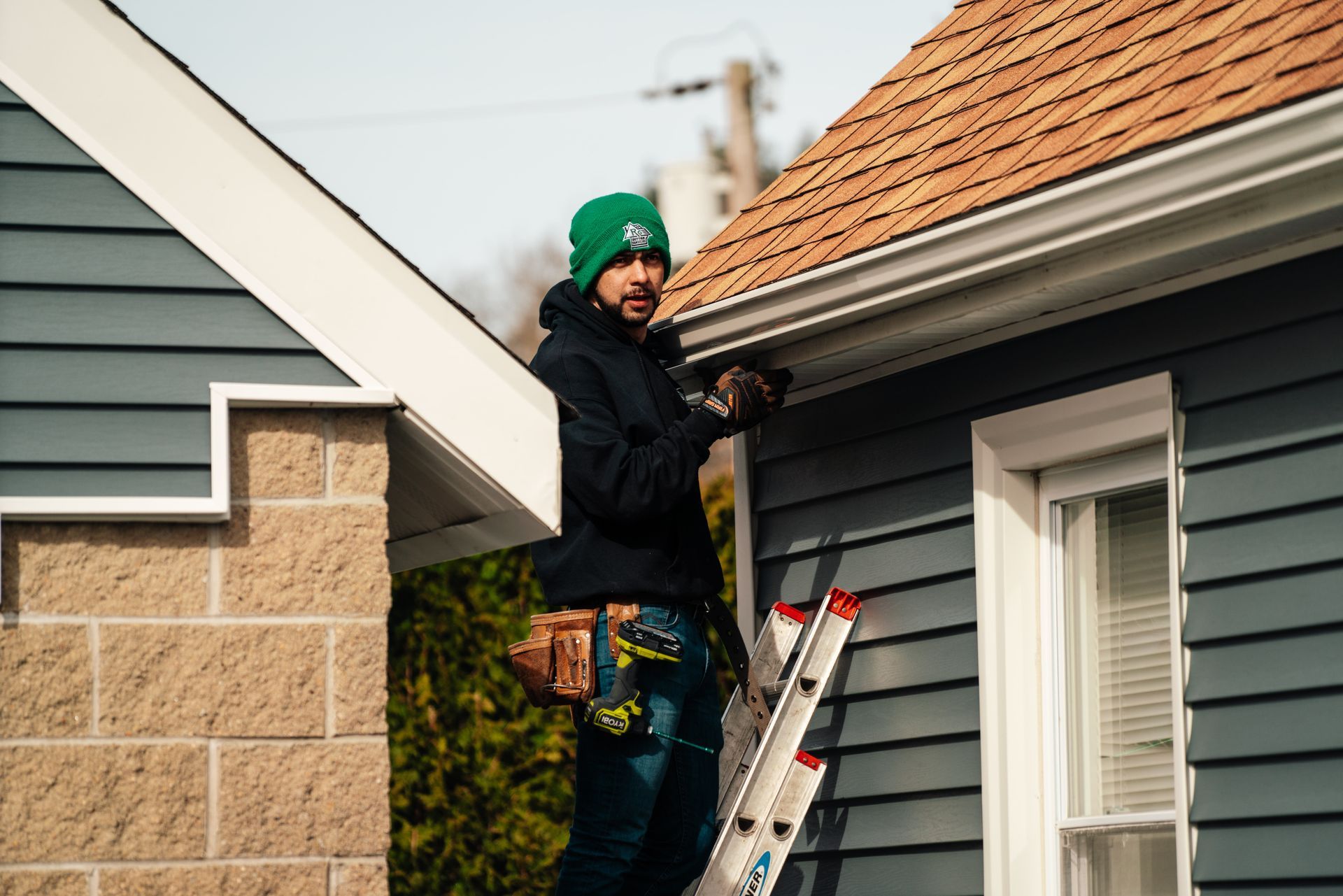 A man is standing on a ladder fixing a gutter on the side of a house.