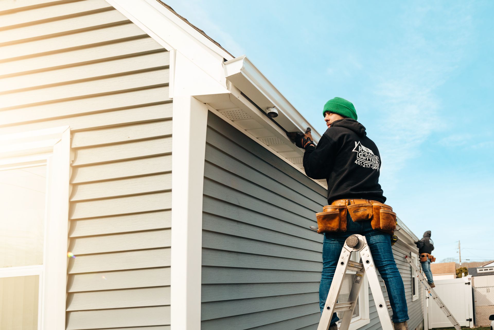 A man is standing on a ladder fixing a gutter on the side of a house.