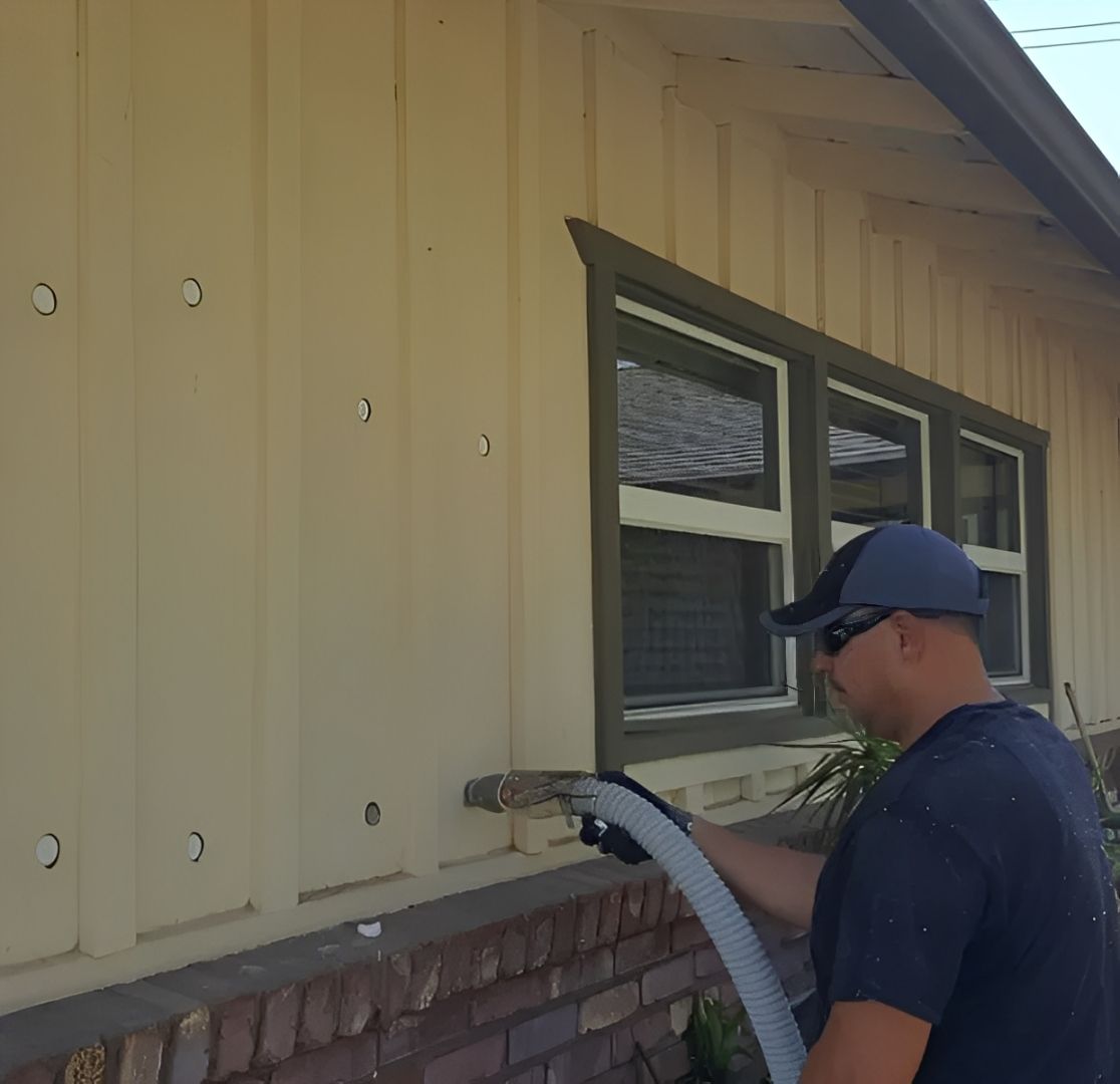 A man is insulating a house wall with a hose. The wall is light yellow with several holes, and the man is wearing a cap and sunglasses.
