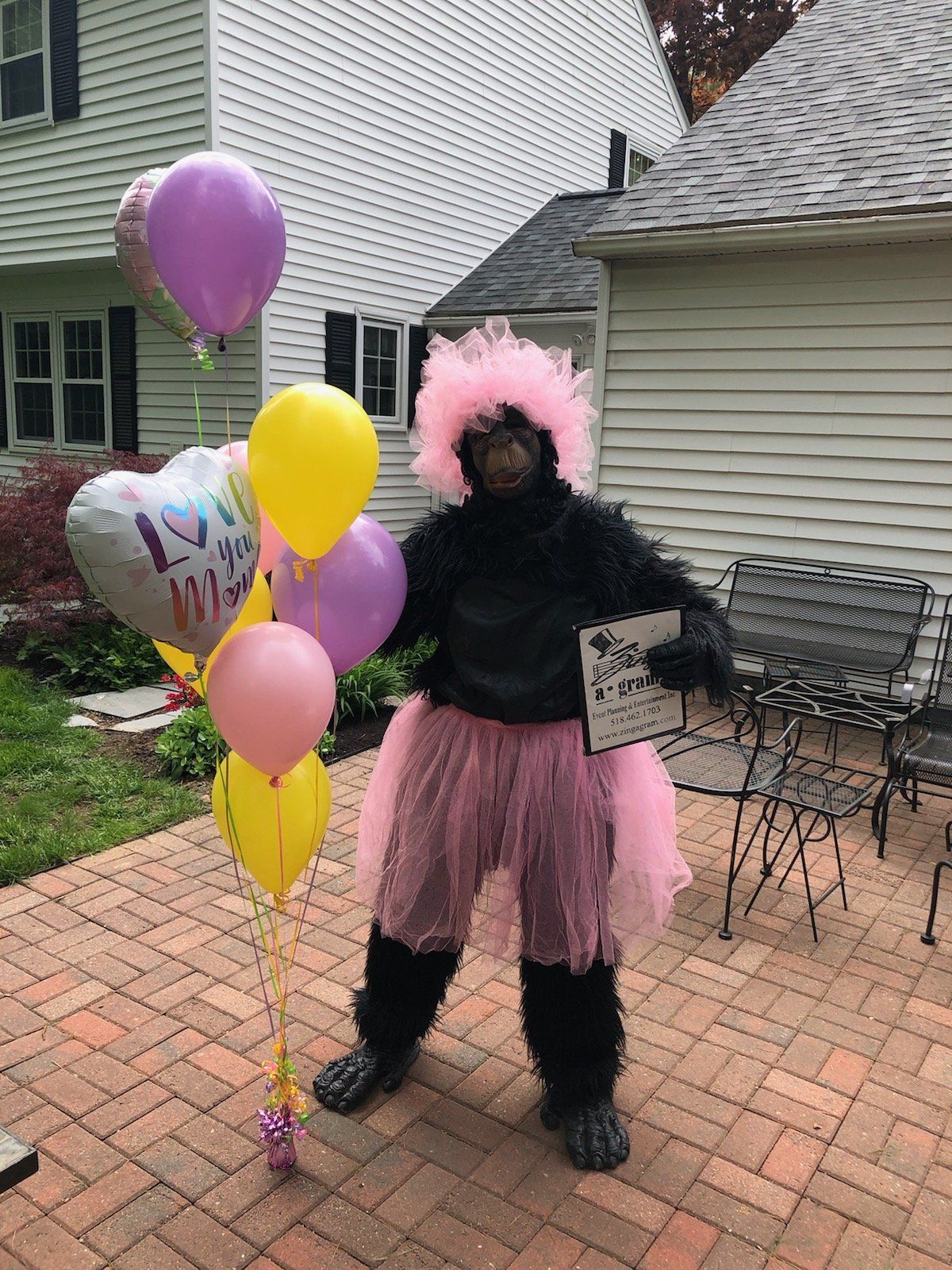 Person in a pink tutu and wig holding balloons and a sign in a brick patio yard.