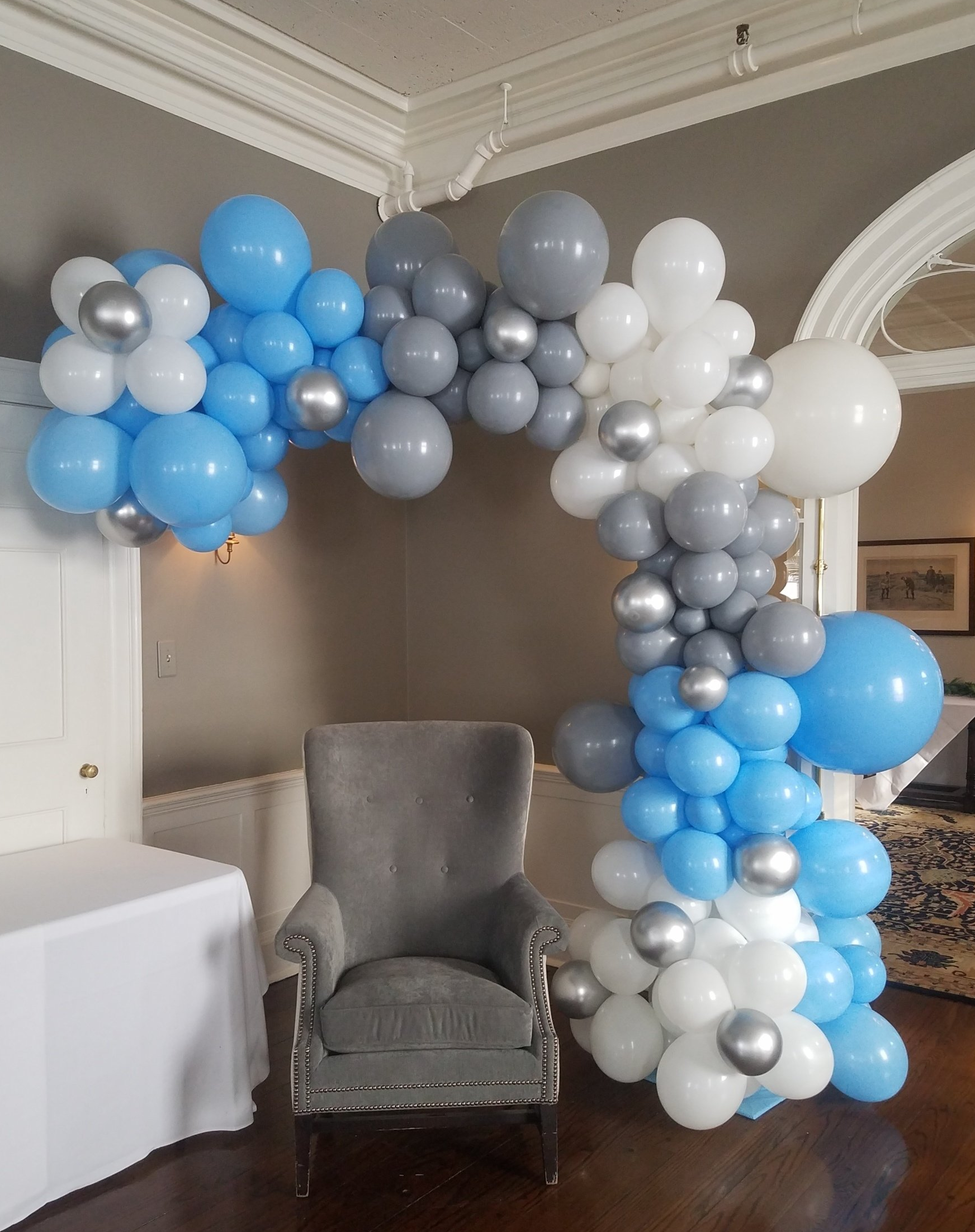 Blue, white, and gray balloon arch framing a gray armchair in an indoor event space