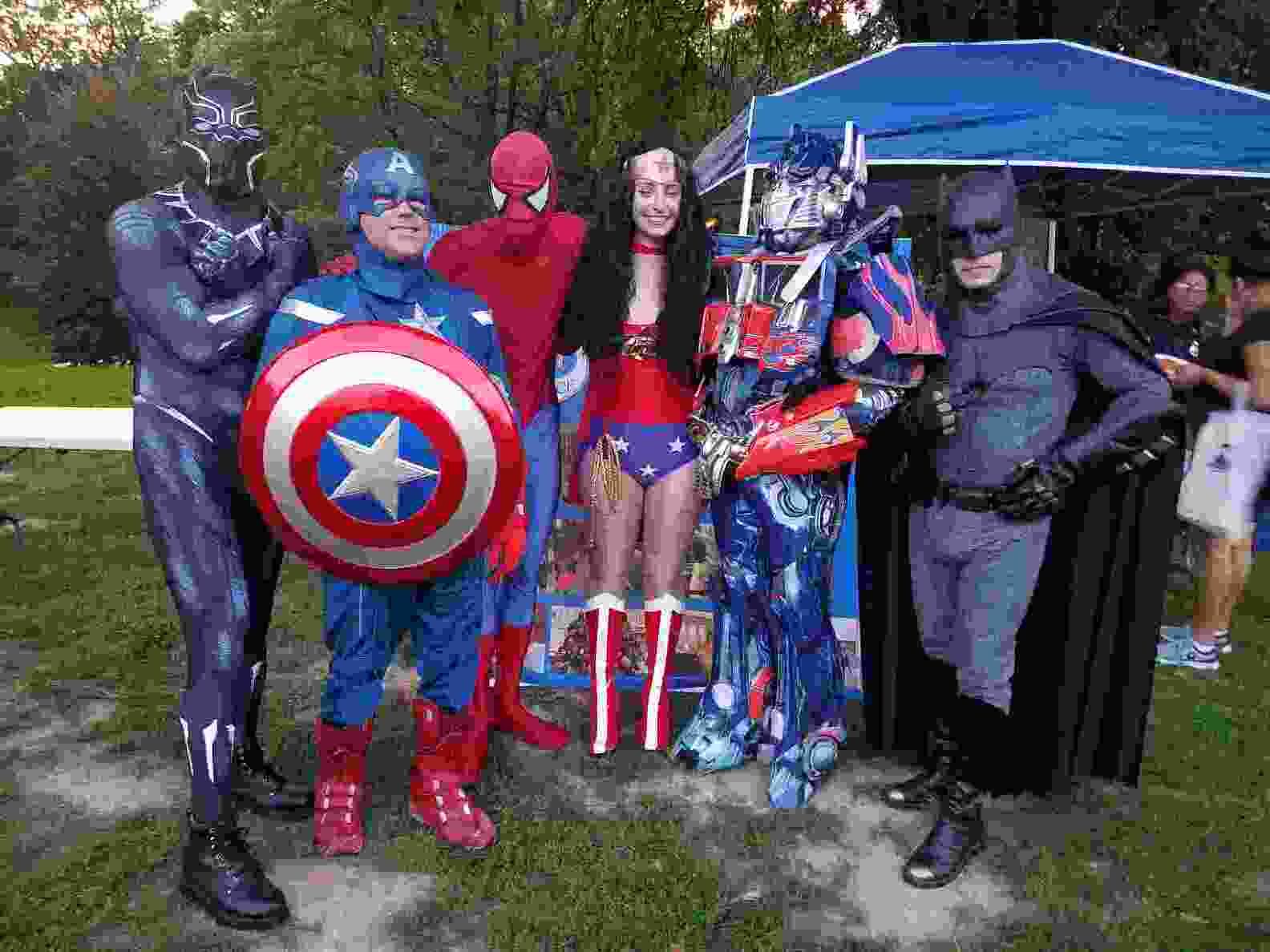 Costumed superheroes posing outdoors in front of trees, including Captain America and Spider-Man, with a child in the center