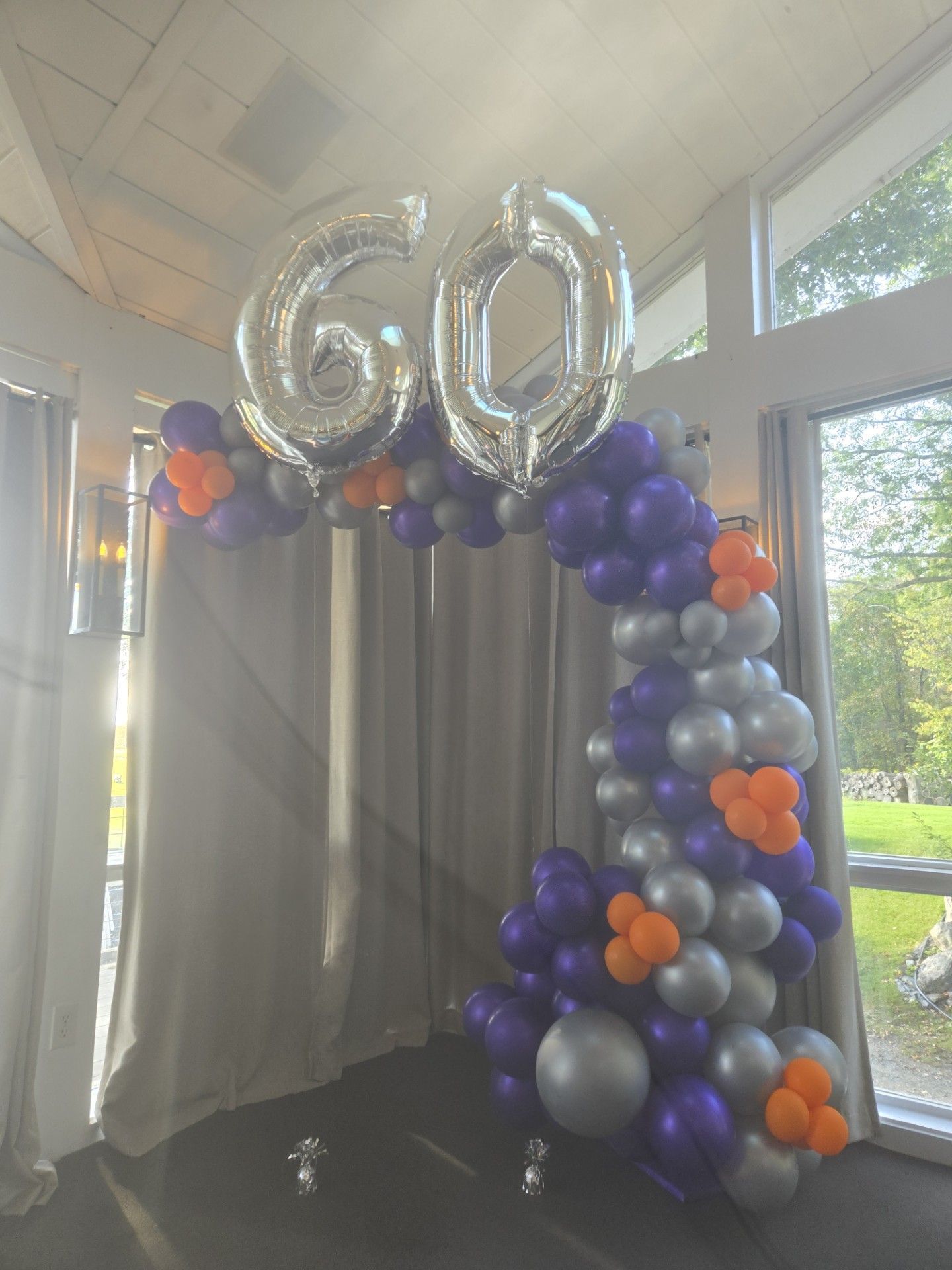 Silver “60” balloons above purple, silver, and orange balloon arch in an indoor celebration setup.