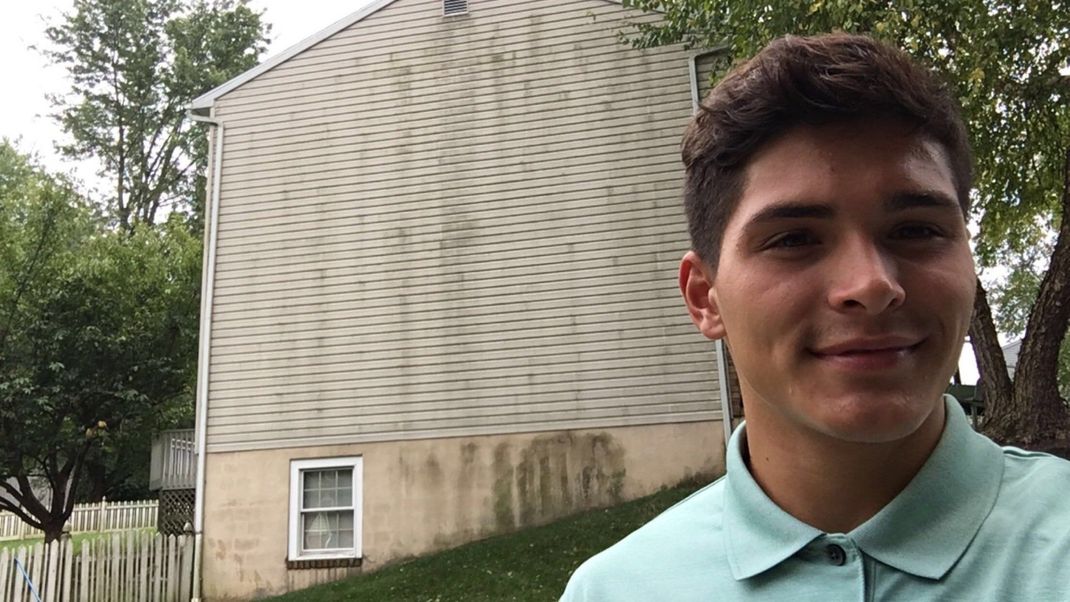 A young man in a blue shirt is standing in front of a house.