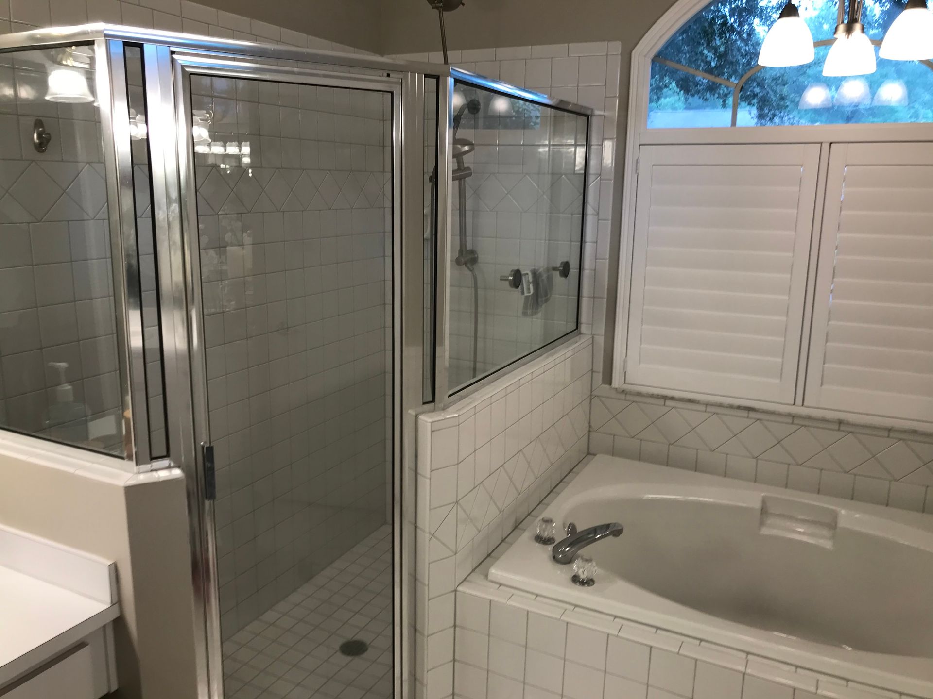 Bathroom with glass shower, bathtub, white tile, and window with shutters.