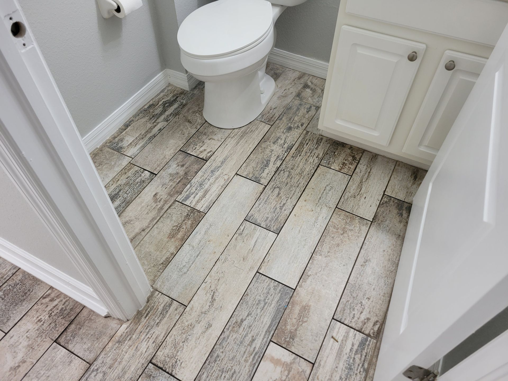 Bathroom with wood-look tile floor, white toilet and cabinets, and a partially open white door.