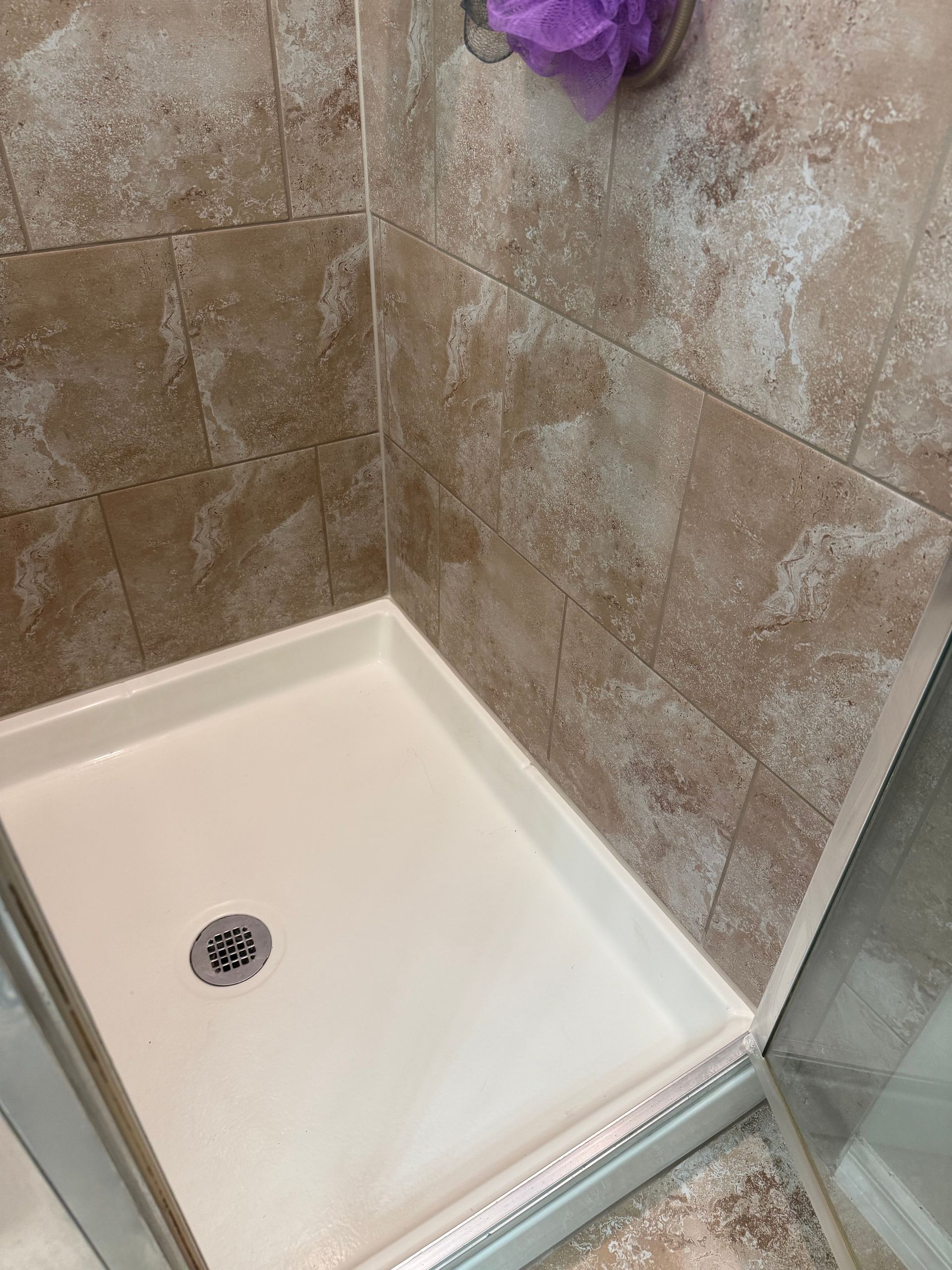 White shower base with brown tile walls, purple loofah, and corner glass door.