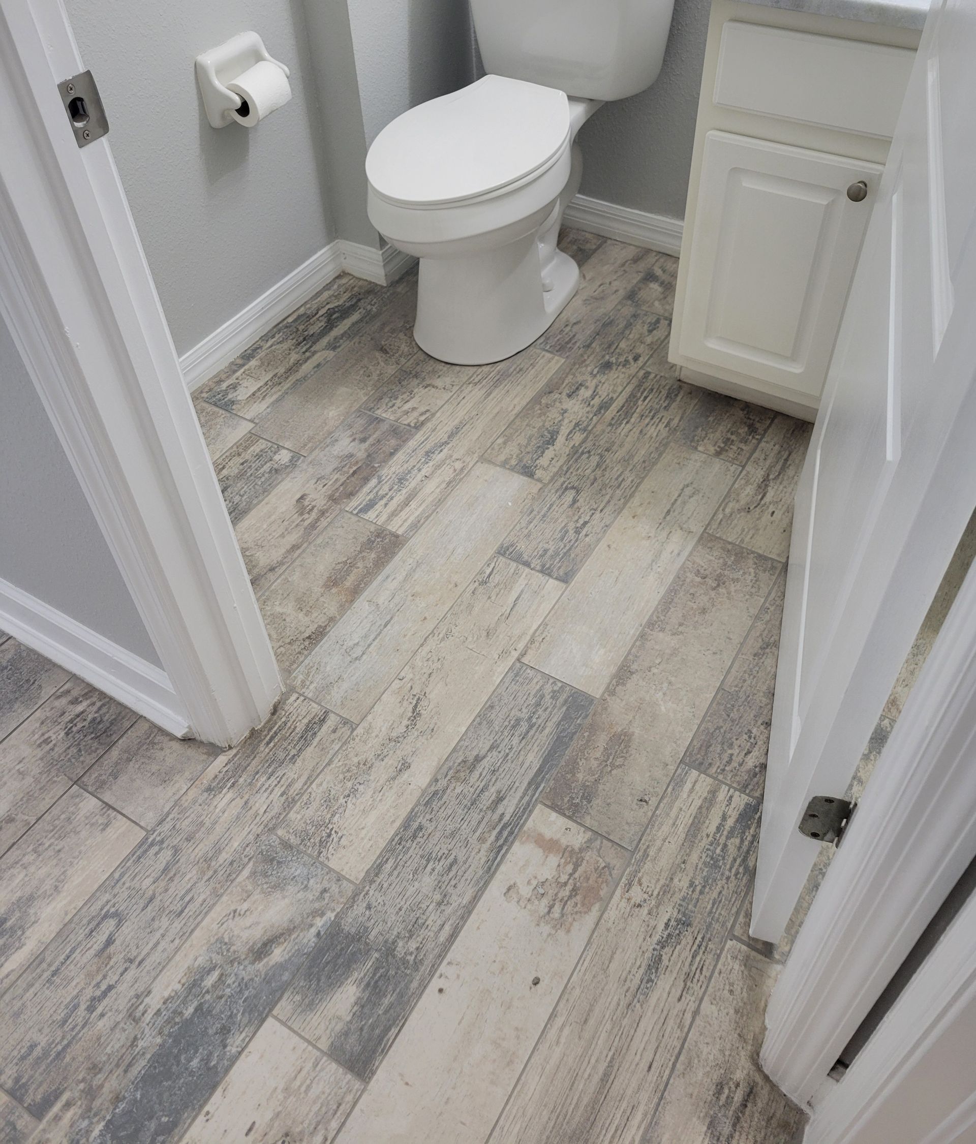 Bathroom with wood-look plank flooring, white toilet, and cabinet. Gray and white tones.