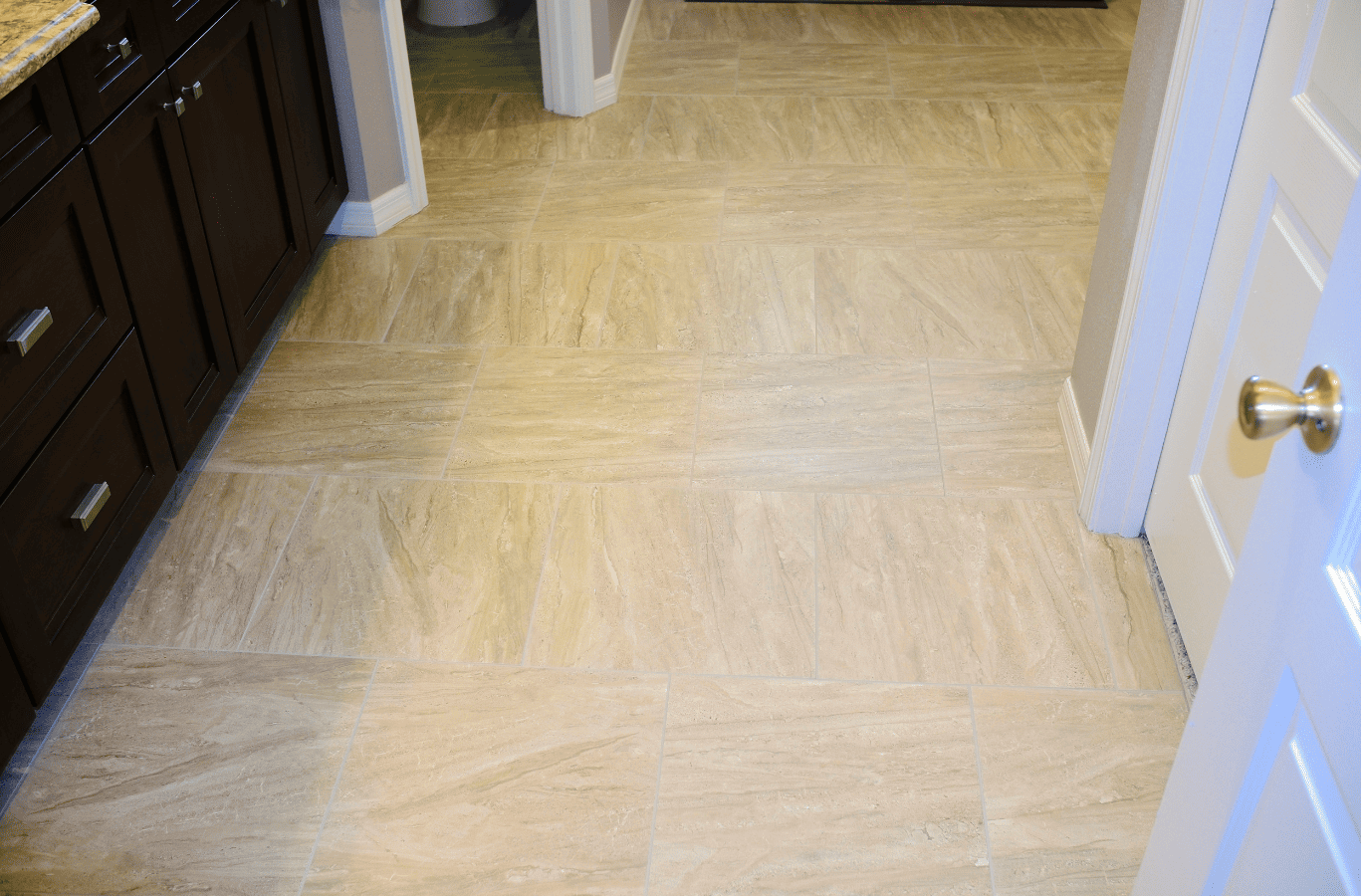 Beige tile floor in a bathroom next to brown cabinets and a white door with a gold doorknob.