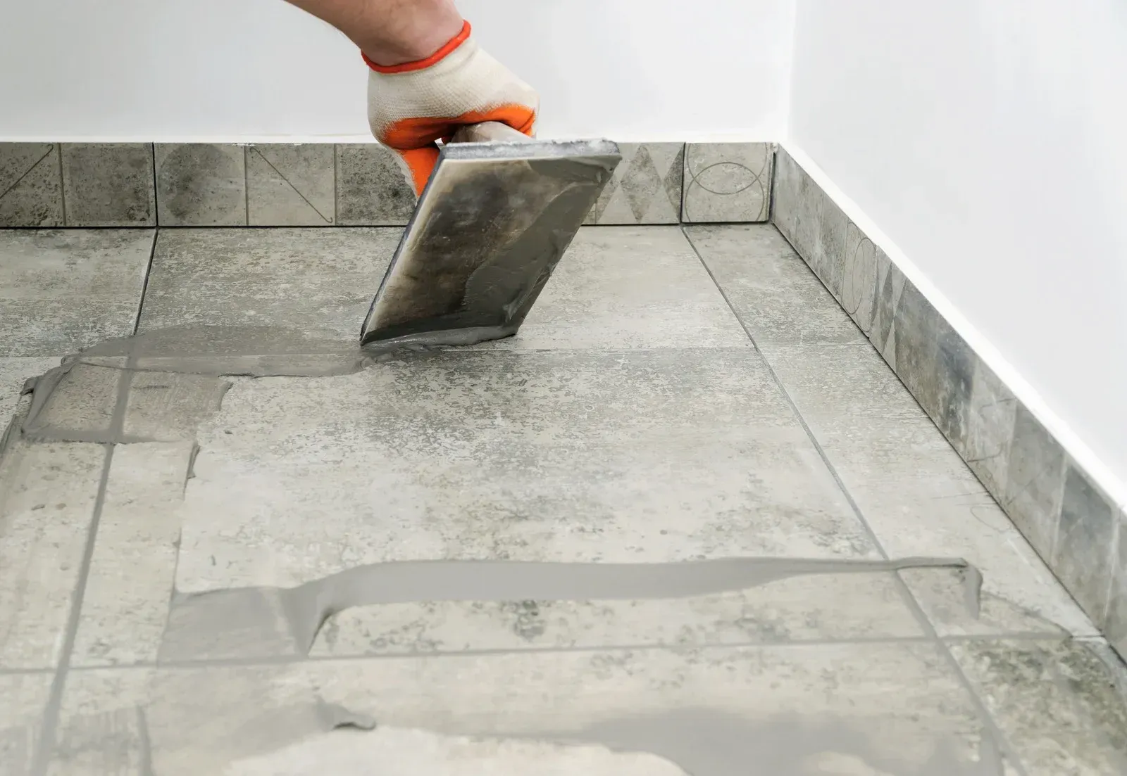 Gloved hand using a trowel to apply grout between gray floor tiles outdoors.