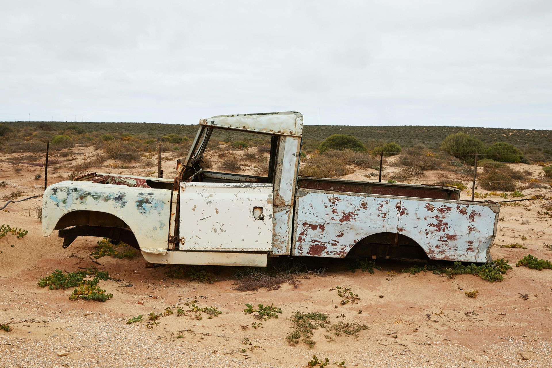 Rusted white and blue pickup truck, missing parts, sits in sandy field under cloudy sky.