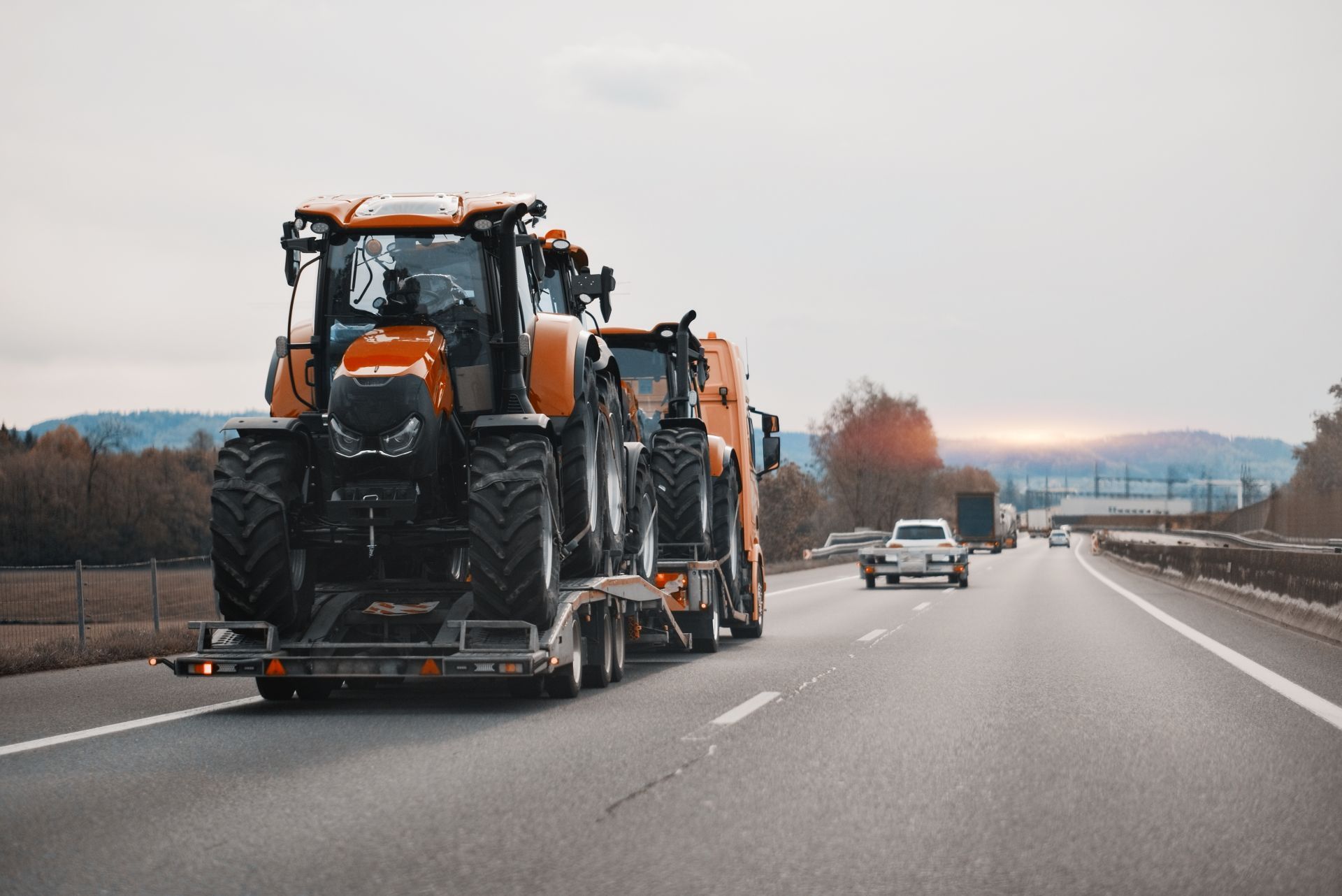 Orange tractors on a flatbed trailer traveling on a highway under a cloudy sky.