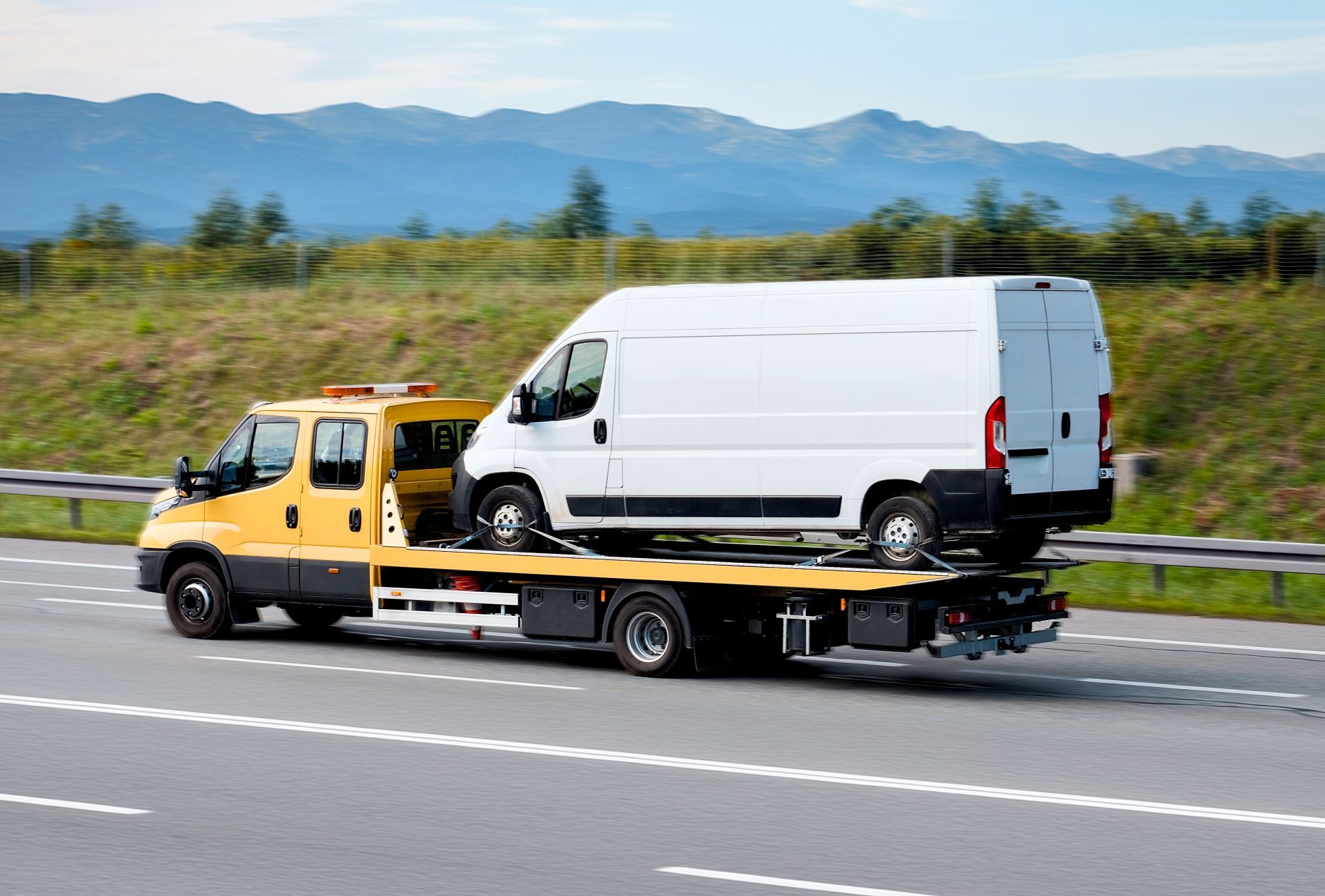Yellow tow truck carrying a white van on a highway, mountains in the background.