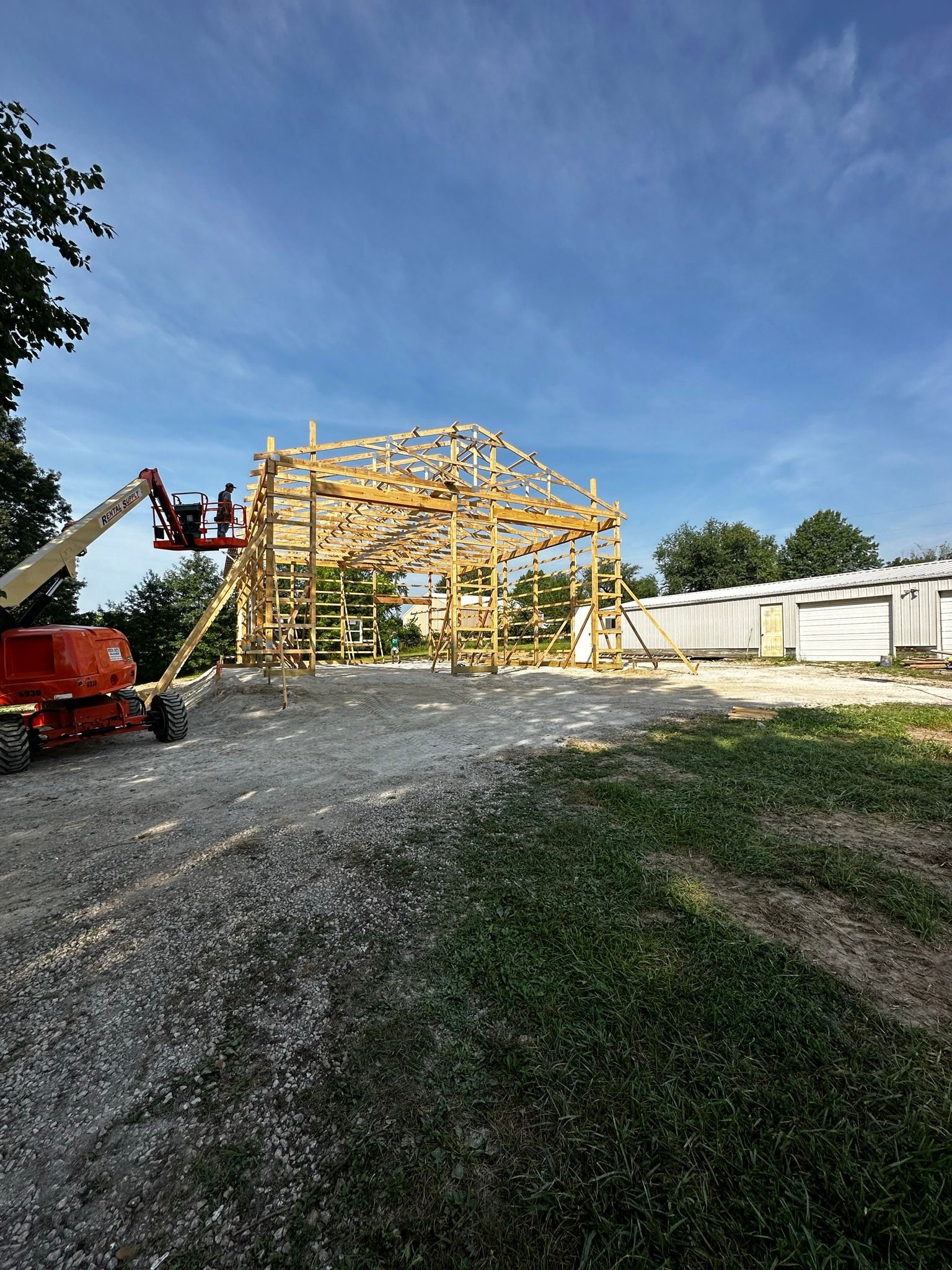 A large wooden post-frame structure is being built in a gravel lot.