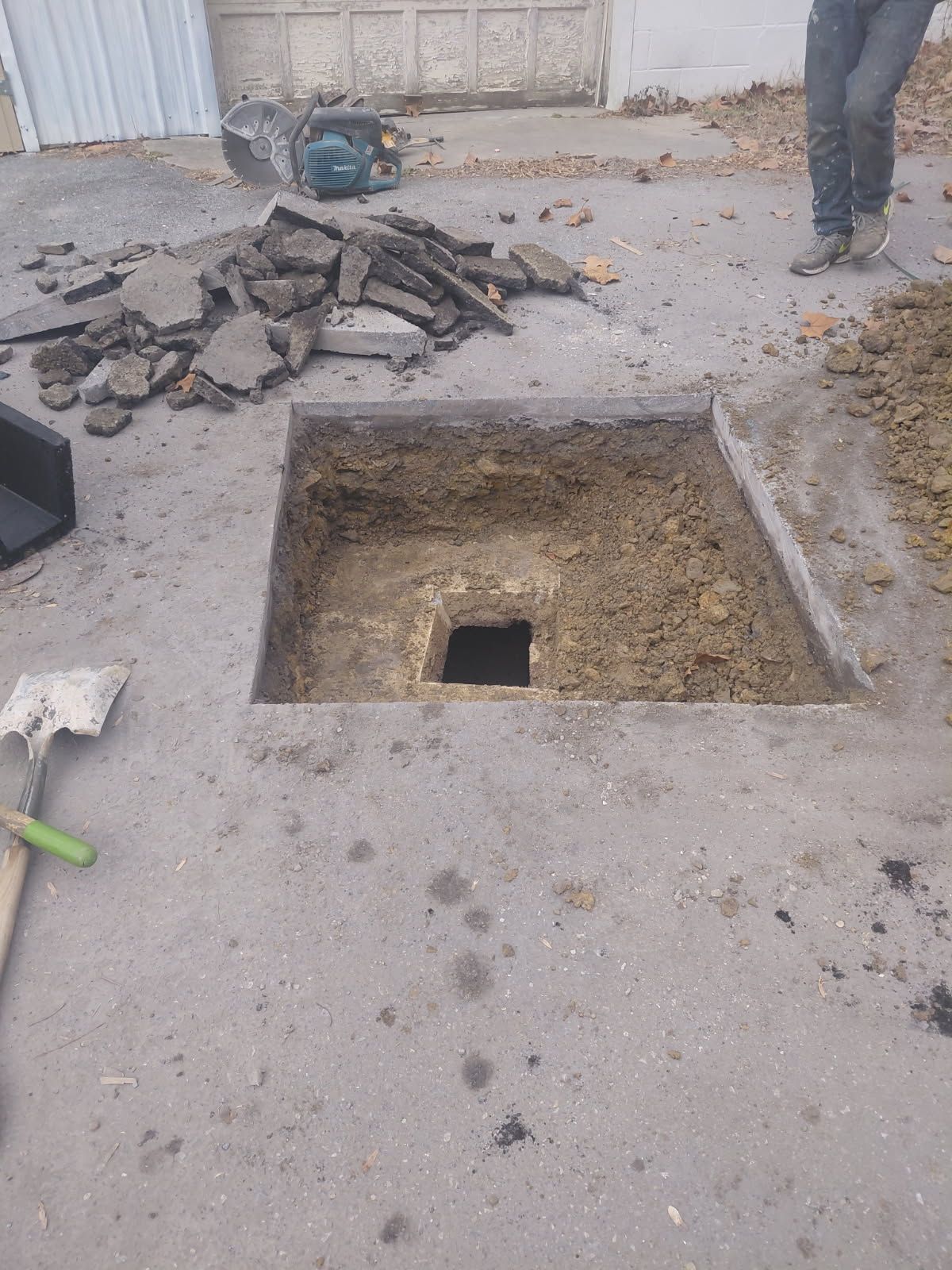 Square hole dug in asphalt. Asphalt debris nearby, person standing. Dirt and tools present.