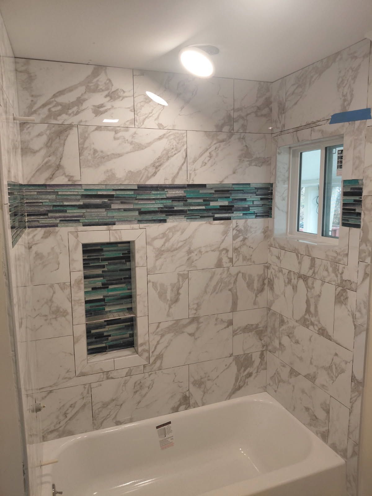 Bathroom with white marble-look tiles, blue-green accent strip, and window above a white tub.