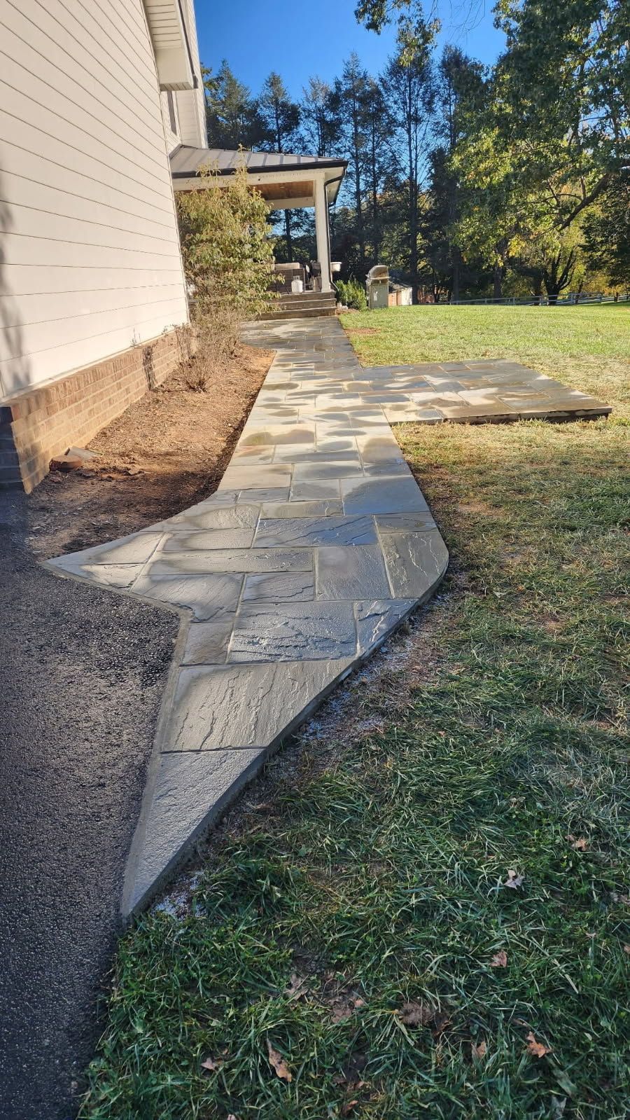 Stone pathway along the side of a white building, leading to a porch and grassy yard on a sunny day.