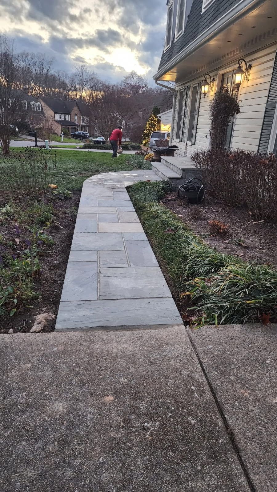 Stone walkway leading to a house with a person in a red jacket, shrubs, and a cloudy sky.
