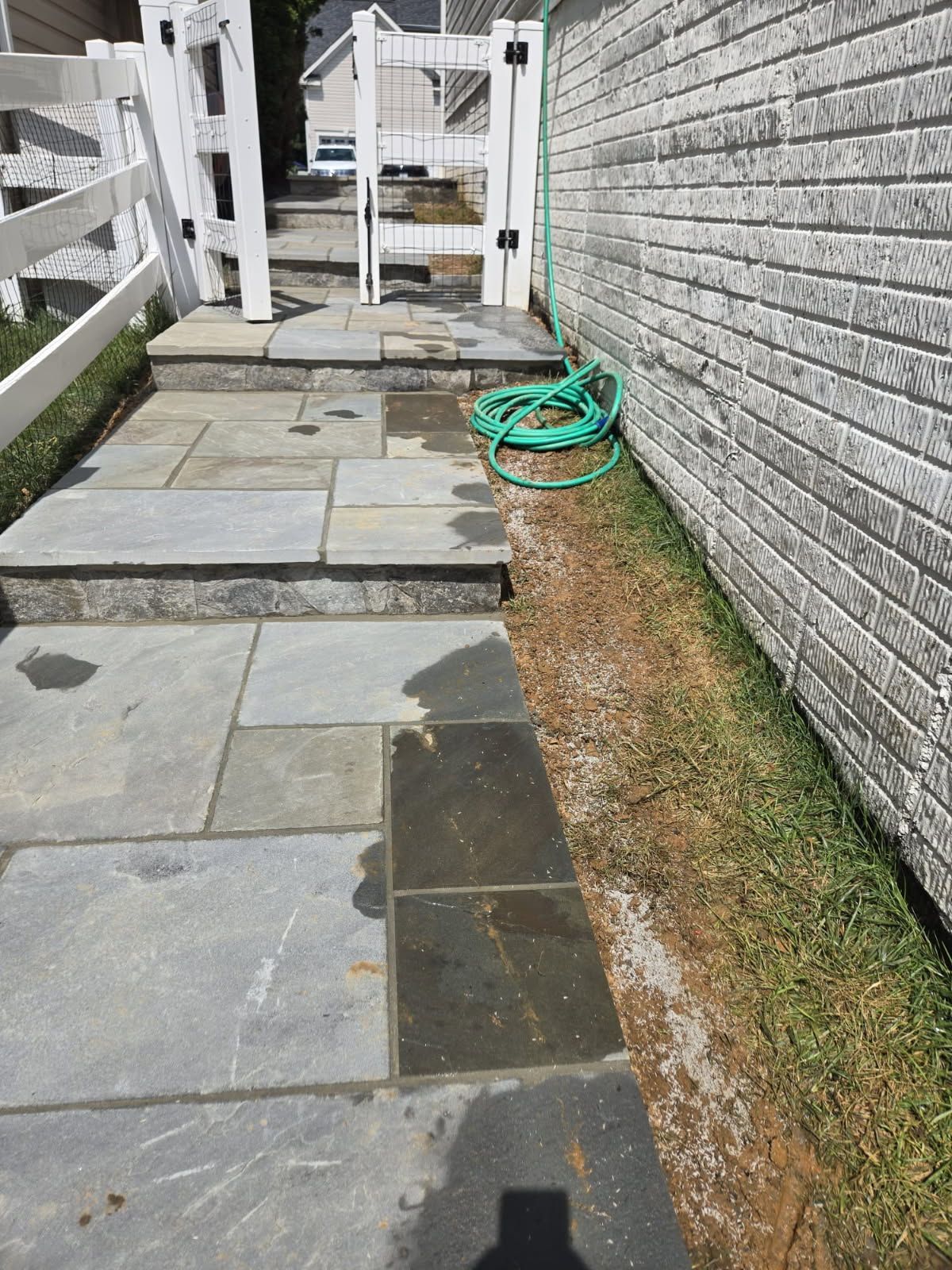 Stone walkway leading to white gate, next to brick wall and grass. A green hose lies on the grass.
