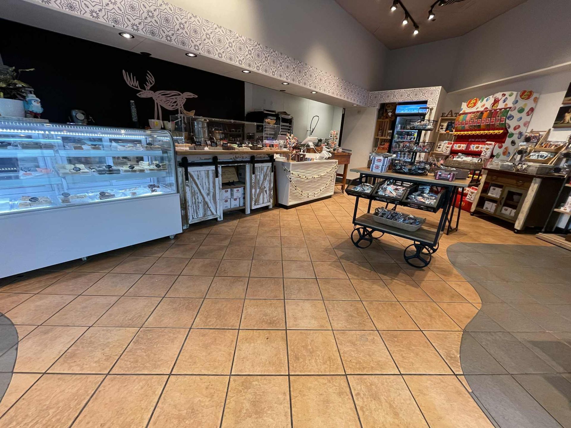 Interior of a chocolate shop with display cases, a cart, and various treats. Beige tile floor, white walls, and dark accents.