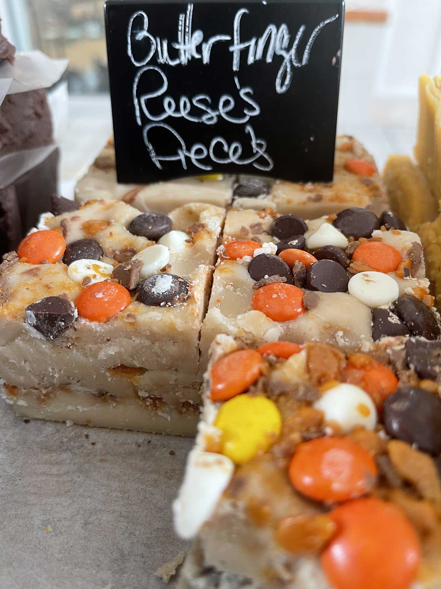 Fudge squares topped with Butterfinger, Reese's Pieces, and chocolate chips, with a handwritten sign.