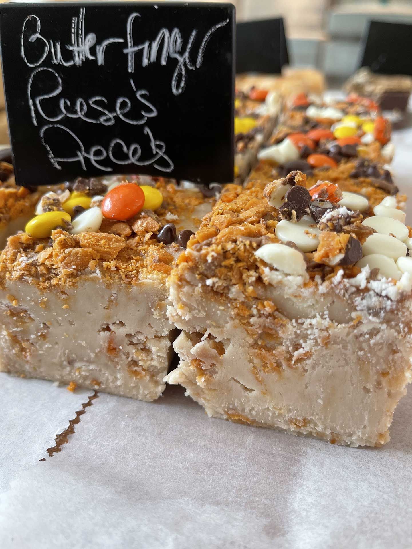 Butterfinger, Reese's Pieces fudge, topped with candies and crumbs, on parchment, with a handwritten sign.