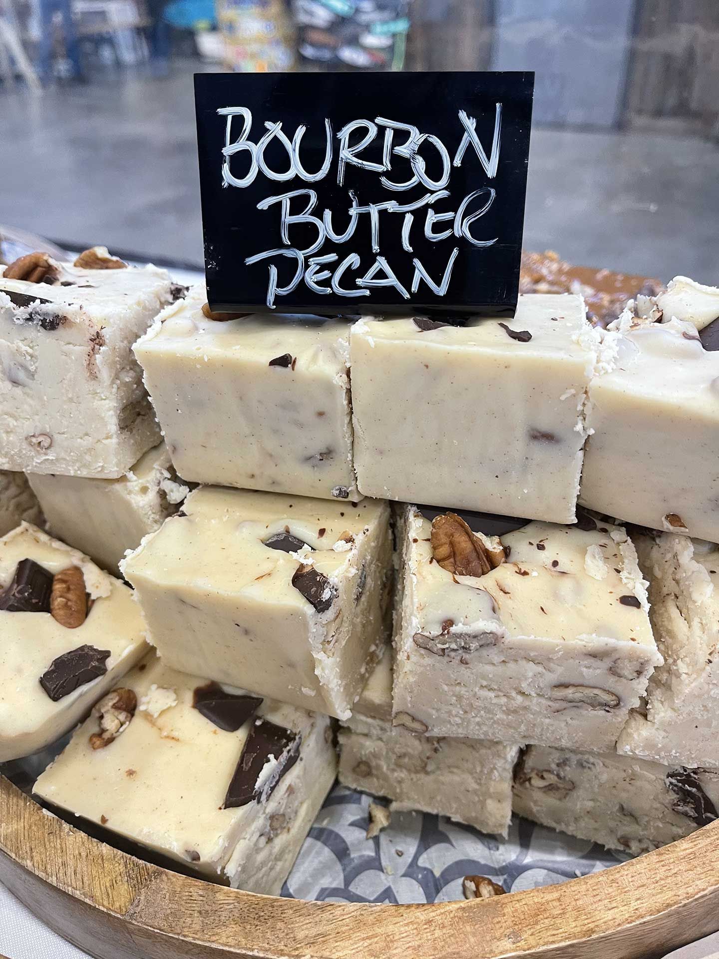 Bourbon butter pecan fudge squares stacked in a wooden tray, labeled with a chalkboard sign.