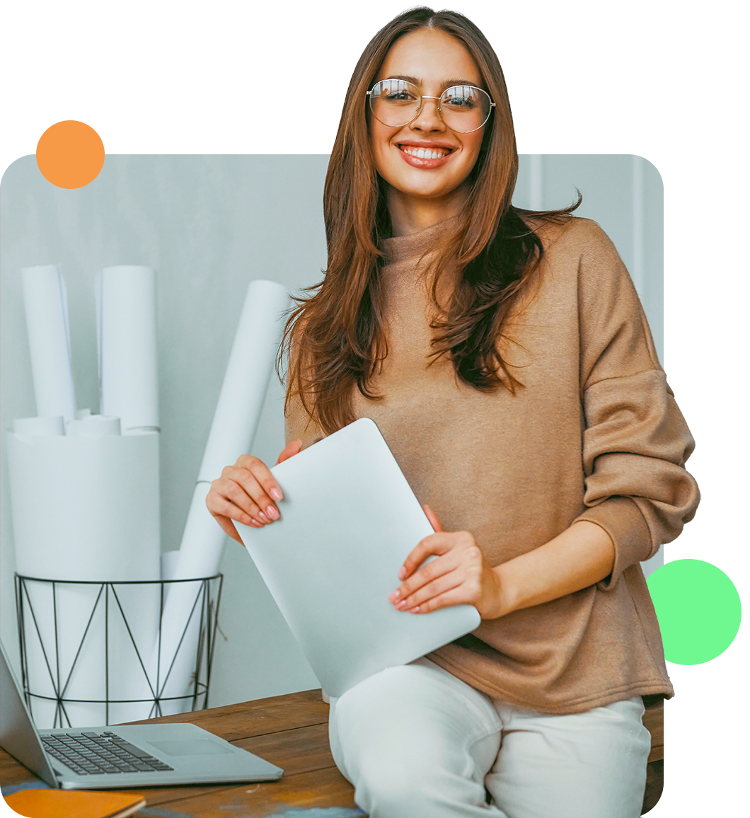 Woman smiling, holding laptop, leaning on desk, with blueprints and computer in office.